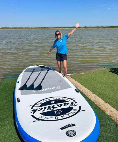 A woman in a blue shirt standing beside the XL Giant FatStick Paddle Board Package on green grass next to a lake, with paddles resting on the board.