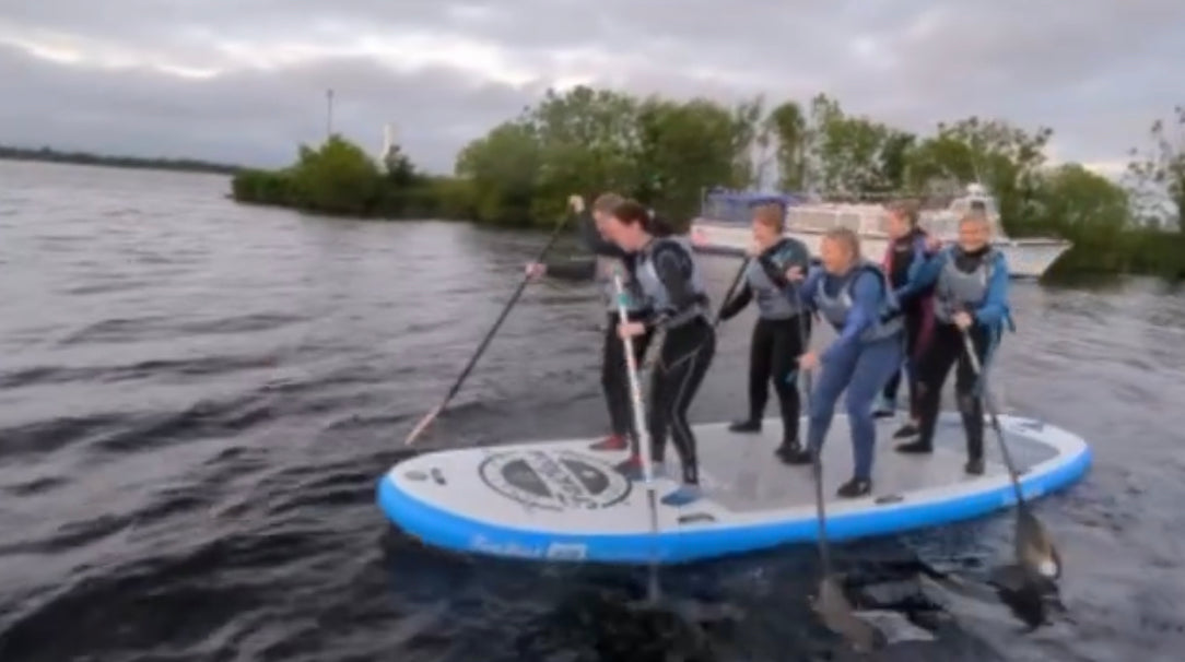 Five people in wetsuits paddling in unison on the XL Giant FatStick Paddle Board Package on dark water with trees in the background.